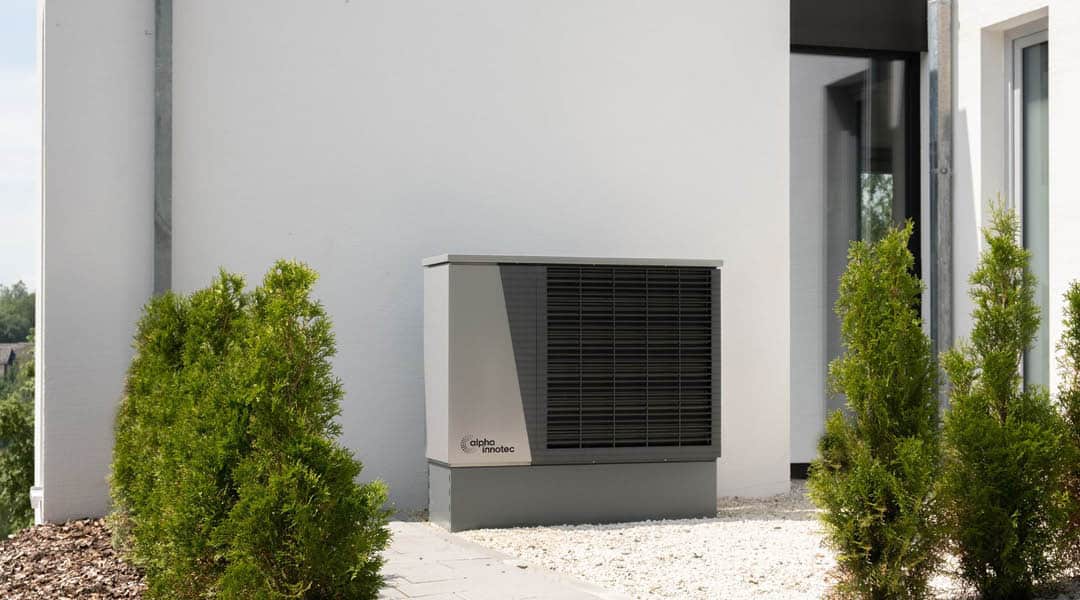 Exterior air‑source heat pump installed beside modern Bespoke Custom Build Home with landscaped gravel and shrubs.